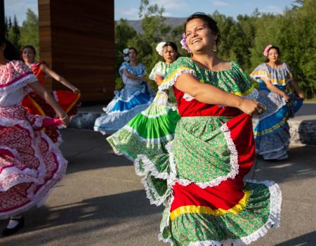 A group of dancers in colorful traditional dresses performs outdoors, led by a smiling woman in a green floral dress with red and yellow accents.